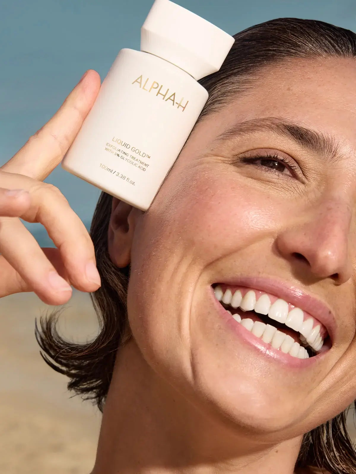 Alpha-H Liquid Gold Exfoliating Treatment 100ml met stralende vrouw die glimlacht en de fles laat zien.