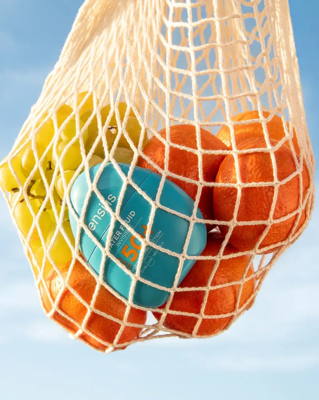 Sensilis Water Fluid 50+ 40ml in mesh bag with oranges and green apples, perfect for summer skin protection.